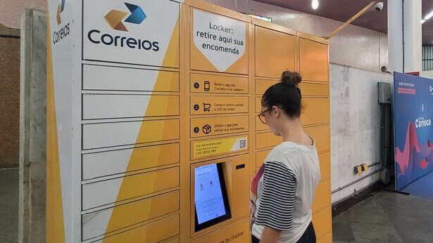 Armário inteligente com a marca dos Correios localizado em uma estação de metrô. O equipamento é composto por uma estrutura metálica, com várias portas individuais. No centro há uma tela de computador com leitor de QR Code para que o sistema seja operado pelo usuário.