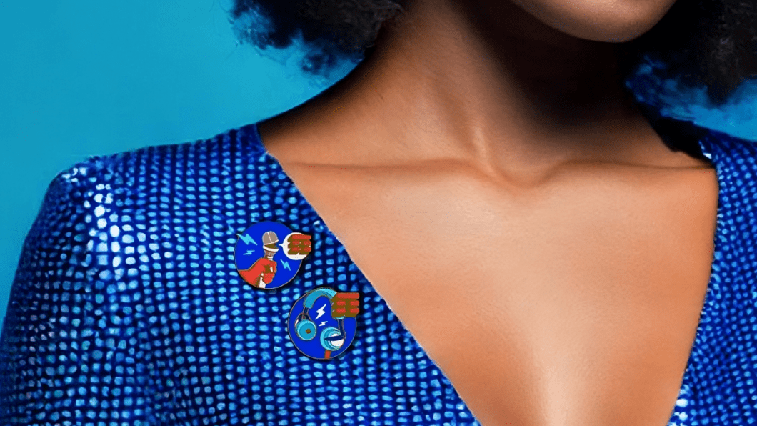 Corpo feminino com uma blusa azul. No lado direito do peito encontram-se dois broches, com ilustrações temáticas de festival e logotipo da TIM.